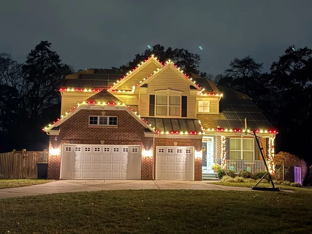 A brick and siding home at dusk with warm white lights along its rooflines and red accents